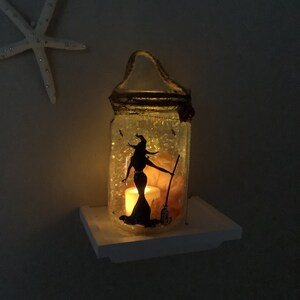 May include: A decorative glass jar with a witch silhouette, lit by a candle. The jar has a rope handle and is decorated with glitter and small embellishments. The witch is holding a broom. The jar is sitting on a white shelf.