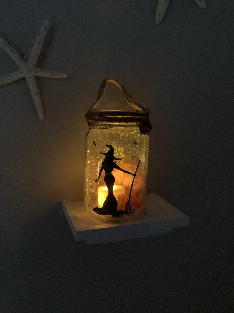 May include: A decorative glass jar with a witch silhouette, lit by a candle. The jar has a rope handle and is decorated with glitter and small embellishments. The witch is holding a broom. The jar is sitting on a white shelf.
