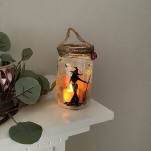 May include: A Halloween-themed mason jar lantern with a witch silhouette, broom, and bats. The jar has a twine handle, a decorative rim, and a small purple flower. A lit candle inside casts a warm glow.