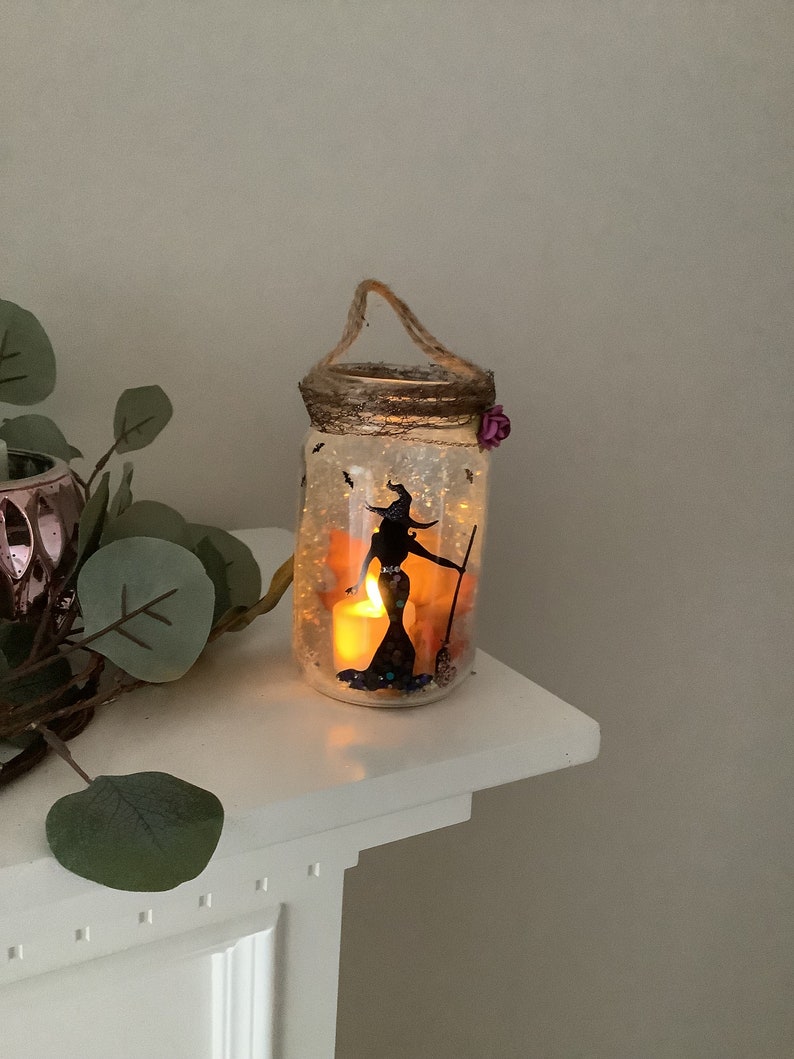May include: A Halloween-themed mason jar lantern with a witch silhouette, broom, and bats. The jar has a twine handle, a decorative rim, and a small purple flower. A lit candle inside casts a warm glow.