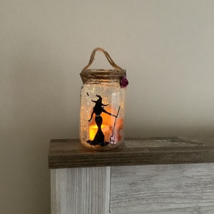 May include: A decorative Halloween-themed jar lantern. The clear glass jar features a black silhouette of a witch holding a broom. The jar is filled with glitter and illuminated by a warm, flickering light. A jute handle and a purple flower add decorative touches.