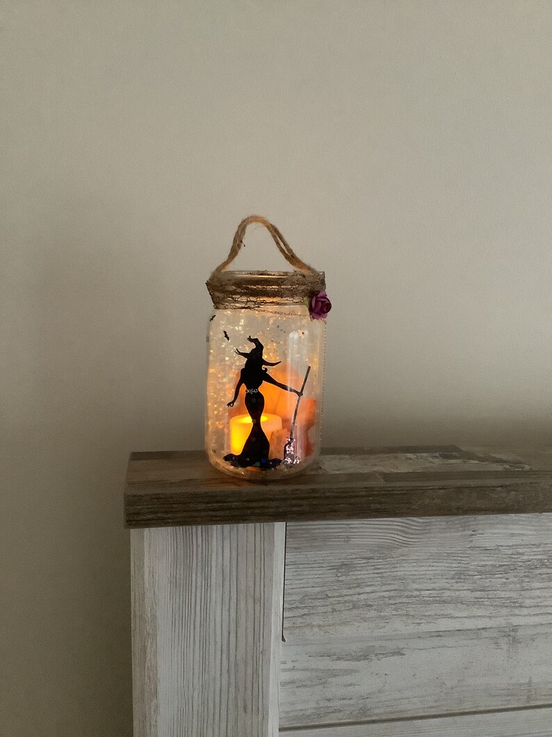 May include: A decorative Halloween-themed jar lantern. The clear glass jar features a black silhouette of a witch holding a broom. The jar is filled with glitter and illuminated by a warm, flickering light. A jute handle and a purple flower add decorative touches.
