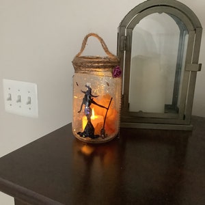 May include: A Halloween-themed mason jar lantern with a witch silhouette, a broom, and a lit candle inside. The jar is decorated with glitter and a twine handle. A decorative metal lantern is next to the jar.
