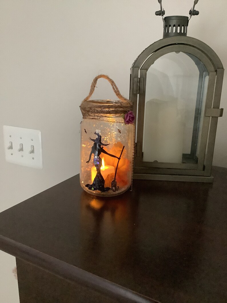 May include: A Halloween-themed mason jar lantern with a witch silhouette, a broom, and a lit candle inside. The jar is decorated with glitter and a twine handle. A decorative metal lantern is next to the jar.