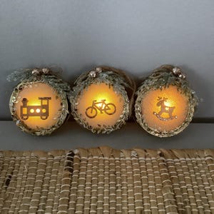 May include: Three illuminated round ornaments with festive designs. Each ornament features a different image: a train, a bicycle, and a rocking horse. The ornaments are trimmed with gold and adorned with glitter and greenery.