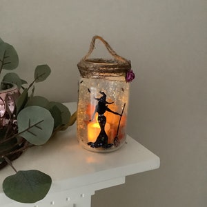 May include: A decorative Halloween-themed jar with a witch silhouette, broom, and a lit candle inside. The jar has a twine handle and a small purple flower. The jar is on a white surface.