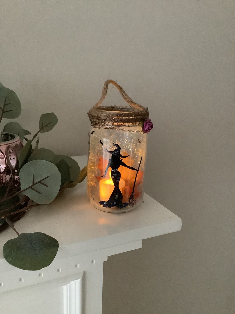 May include: A decorative Halloween-themed jar with a witch silhouette, broom, and a lit candle inside. The jar has a twine handle and a small purple flower. The jar is on a white surface.