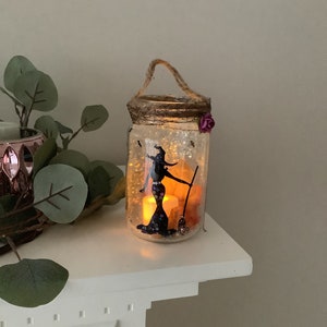 May include: A decorative Halloween-themed jar with a witch silhouette, broom, and glowing lights. The jar has a rope handle and a decorative rim with a purple flower. The jar is filled with decorative elements and lights.