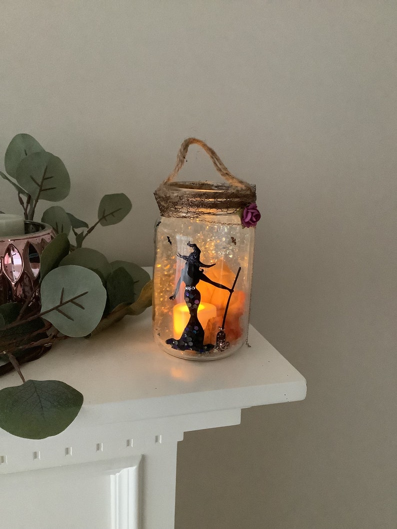 May include: A decorative Halloween-themed jar with a witch silhouette, broom, and glowing lights. The jar has a rope handle and a decorative rim with a purple flower. The jar is filled with decorative elements and lights.