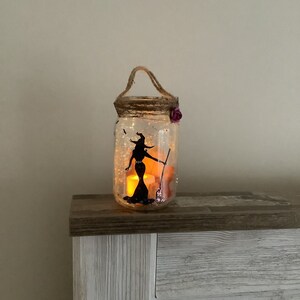 May include: A decorative Halloween-themed mason jar lantern. The clear glass jar features a black silhouette of a witch holding a broom. The jar is decorated with glitter and a purple flower. A rope handle is attached to the top.