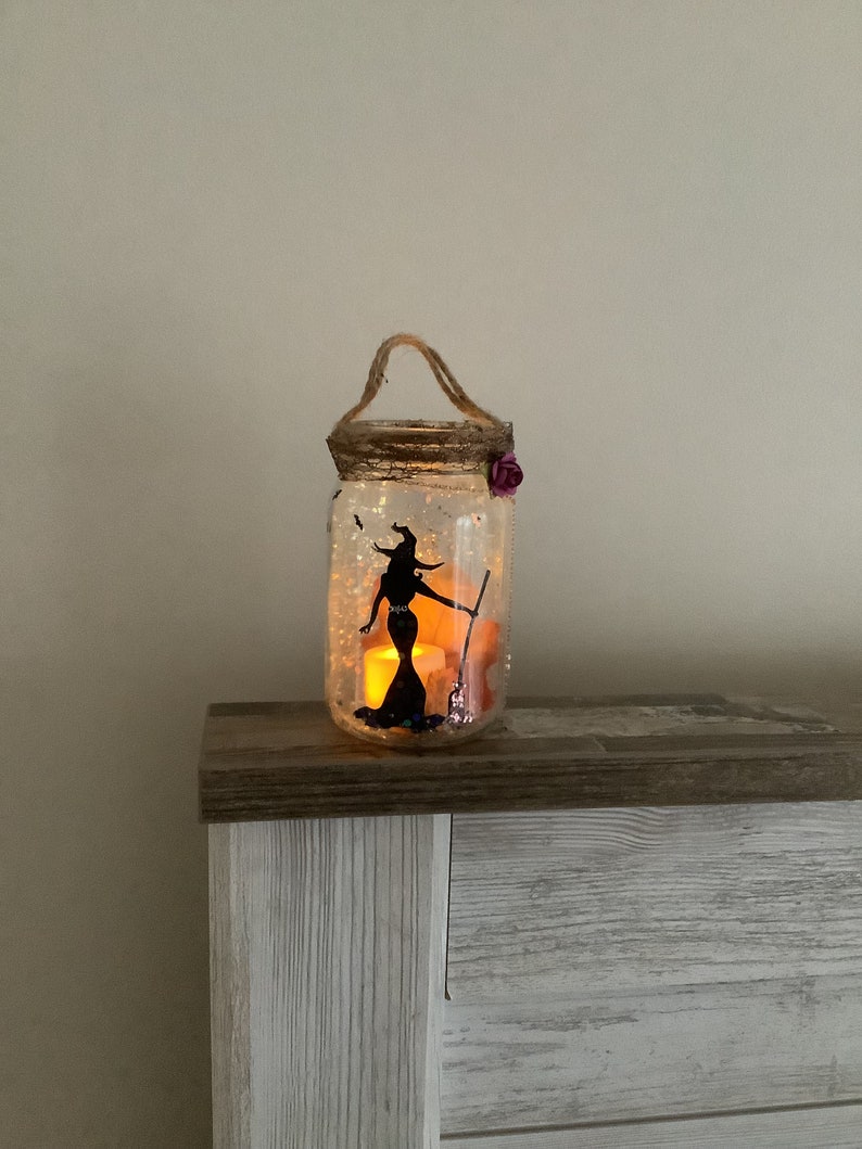 May include: A decorative Halloween-themed mason jar lantern. The clear glass jar features a black silhouette of a witch holding a broom. The jar is decorated with glitter and a purple flower. A rope handle is attached to the top.