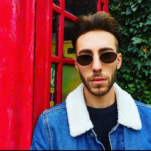 Puede incluir: Un hombre con una chaqueta vaquera azul con cuello de borreguito blanco y gafas de sol ovaladas. Está de pie frente a una cabina telefónica roja.
