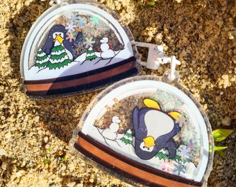 May include: Two clear acrylic snow globe keychains. Each keychain features a penguin, snowman, and evergreen trees on a snowy landscape. The keychains have a brown border and are filled with glitter.