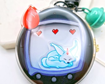 May include: A black and clear plastic keychain shaped like a Tamagotchi with a blue screen displaying a white and blue sleeping fox with a red horn. The keychain has a red heart charm and a green bell charm.