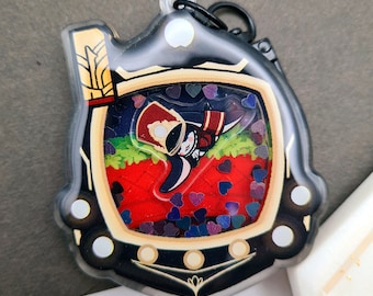 May include: A black and gold charm with a clear plastic cover. Inside the cover is a small cartoon character wearing a red hat and a black and white uniform. The character is surrounded by red and purple glitter hearts.