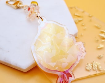 May include: A clear acrylic keychain in the shape of a chick, with a gold-colored clasp and chain. The chick has a yellow, faceted design and pink and red accents. The keychain is on a white and clear surface.