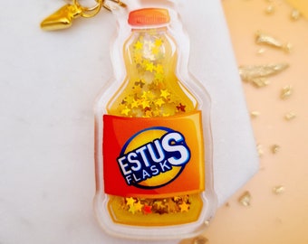 May include: A clear plastic keychain shaped like a bottle with a gold glitter filling and the text "ESTUS FLASK" on the label. The keychain has a gold metal ring and a gold heart charm.