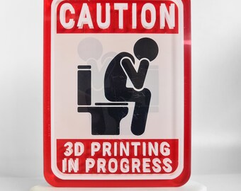 May include: A rectangular sign with a red border and the word "CAUTION" in white. The sign features a black silhouette of a person sitting on a toilet, with the text "3D PRINTING IN PROGRESS" below.