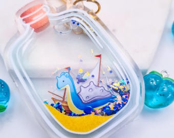 May include: Clear, bottle-shaped keychain with a blue and yellow sea creature design inside. The keychain features a small boat, a red flag, and blue glitter. The bottle is filled with a clear liquid and has a gold-colored keyring.
