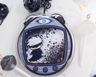 May include: A clear acrylic keychain featuring a cartoon character inside a retro-style device. The keychain has a silver border and a black chain. The character is wearing a blindfold and black clothing. The background has black and white speckles.