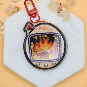 Può includere: Un portachiavi rotondo e trasparente con chiusura rossa. Il portachiavi presenta un personaggio dei cartoni animati in una scena infuocata con accenti di stelle dorate. Il bordo è nero con un motivo a mattoni.