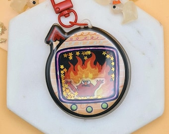 May include: A clear, round keychain with a red clasp. The keychain features a cartoon character in a fiery scene with gold star accents. The border is black with a brick pattern.