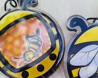 May include: Two bee-themed keychains. One keychain features a cartoon bee inside a honeycomb design, with yellow and black accents. The other keychain has a bee wing design with yellow and black accents. Both keychains have a clear, glossy finish.