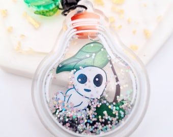 May include: A clear, lightbulb-shaped keychain with a cartoon character inside. The character is white with a black face and is surrounded by green leaves and glitter. The keychain has a black clasp and a green leaf charm.