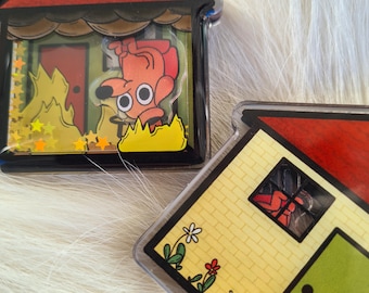 May include: Two house-shaped keychains. One keychain features a cartoon dog in a burning house with yellow flames and star-shaped glitter. The other keychain has a yellow brick house with a red roof and a green door.