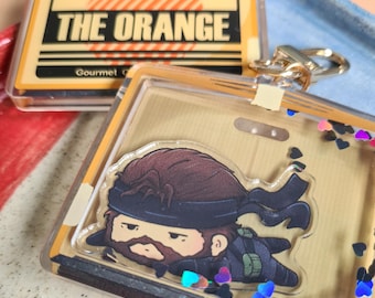 May include: A clear acrylic keychain featuring a cartoon character with a brown beard and a black headband. The keychain has a gold-colored clasp and is filled with heart-shaped confetti. The text "THE ORANGE" is visible on a separate tag.