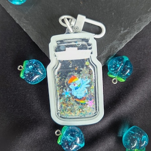 Op de afbeelding: Een doorzichtige sleutelhanger in de vorm van een pot met een stripfiguur erin, omringd door glitter. De sleutelhanger heeft een zilveren sluiting. Er liggen verschillende blauwe, aardbeivormige bedels met groene steeltjes verspreid.