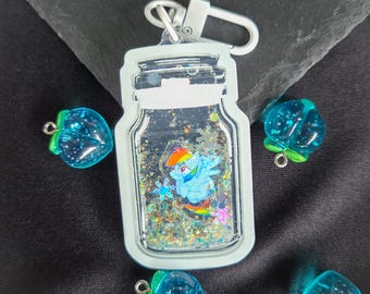 May include: A clear jar-shaped keychain with a cartoon character inside, surrounded by glitter. The keychain has a silver clasp. Scattered around the keychain are several blue, strawberry-shaped charms with green stems.