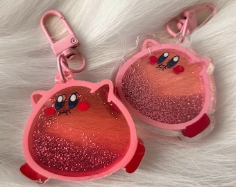 May include: Two pink keychains featuring a cartoon character design. The character is pink with red accents, white eyes, and a smiling expression. The keychains have a glittery interior and a pink clasp.