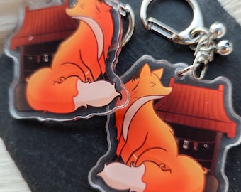 May include: Two clear acrylic keychains featuring an orange fox with a white-tipped tail, sitting in front of a red-roofed structure. Each keychain has a silver-colored clasp. The keychains are on a dark surface.