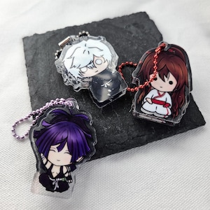 May include: Three acrylic keychains featuring chibi-style anime characters. One has white hair and a black outfit, one has long reddish-brown hair in a white and red outfit with a red chain, and one has purple hair in a black outfit with a purple chain.
