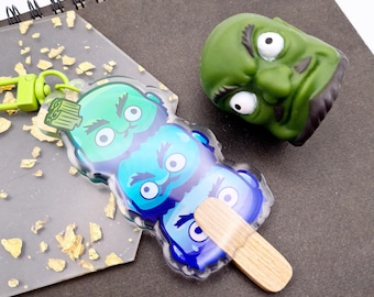 May include: A green, blue, and white cartoon character keychain with a wooden stick, and a green rubber toy head with white eyes and black hair, resting on a black spiral notebook.