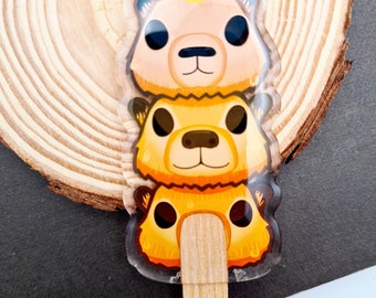May include: A keychain featuring three stacked animal faces, a llama, a fox, and a bear, with a wooden stick attached. The keychain is made of clear acrylic and has a yellow, orange, and brown color scheme.