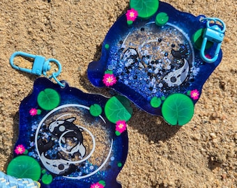 May include: Two blue resin keychains with a koi fish design. The keychains feature a dark blue border with glitter, green lily pads, and pink flowers. Each keychain has a light blue clasp.