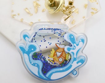 May include: A clear plastic keychain with a gold bell charm. The keychain features a cartoon goldfish swimming in a blue and white wave design with blue glitter.