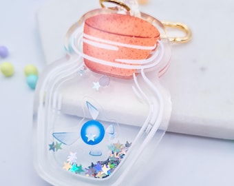 May include: A clear acrylic keychain shaped like a jar with a cork lid. Inside the jar is a blue and white fairy with iridescent glitter. The keychain has a gold metal ring.