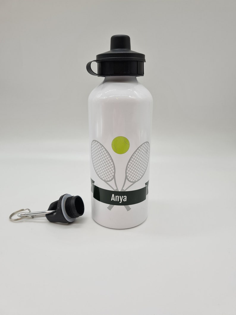 May include: White aluminum water bottle with a black lid and a black carrying loop. The bottle has a tennis racket and ball design with the name "Anya" printed on a black banner.