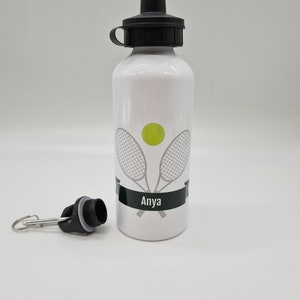 May include: White aluminum water bottle with a black lid and a black carrying strap. The bottle has a tennis racket and ball design with the name "Anya" printed on it.