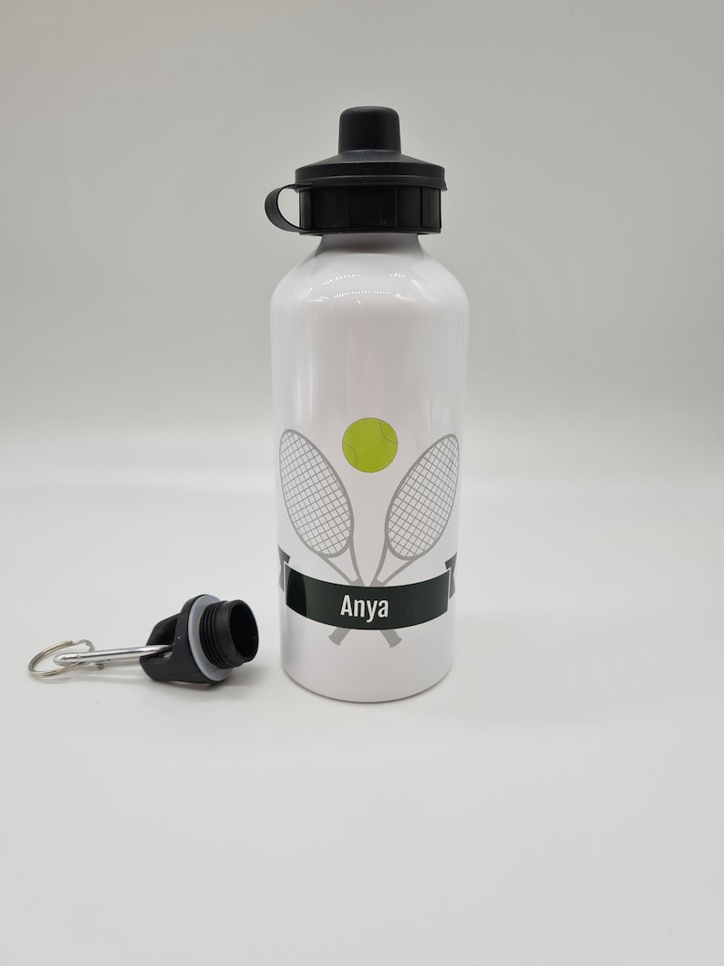 May include: White aluminum water bottle with a black lid and a black carrying strap. The bottle has a tennis racket and ball design with the name "Anya" printed on it.
