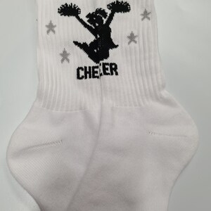 May include: White crew socks with a black silhouette of a cheerleader holding pom-poms and the word "CHEER" printed on the front. The socks have gray stars printed on the sides.
