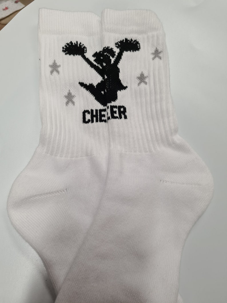 May include: White crew socks with a black silhouette of a cheerleader holding pom-poms and the word "CHEER" printed on the front. The socks have gray stars printed on the sides.