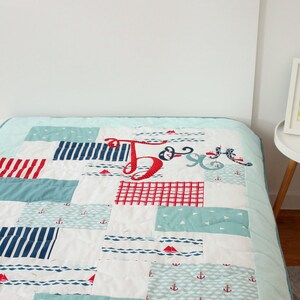 May include: A blue and white patchwork quilt with red accents. The quilt features a nautical theme with anchors, sailboats, and waves. The quilt is made of various fabrics and has a quilted texture.