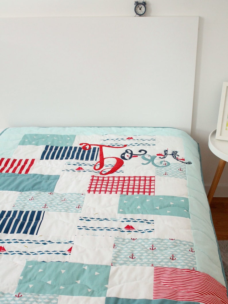 May include: A blue and white patchwork quilt with red accents. The quilt features a nautical theme with anchors, sailboats, and waves. The quilt is made of various fabrics and has a quilted texture.