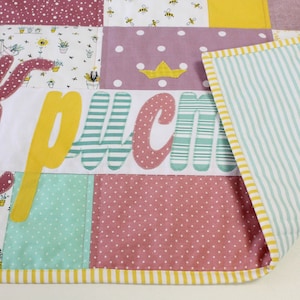 Könnte beinhalten: Ein Patchwork-Quilt mit verschiedenen gemusterten Quadraten in Rosa, Gelb und Mintgrün. Der Quilt zeigt den Namen "Kрисм" in großen, farbenfrohen Buchstaben. Die Rückseite hat ein mintgrünes und weiß gestreiftes Muster.