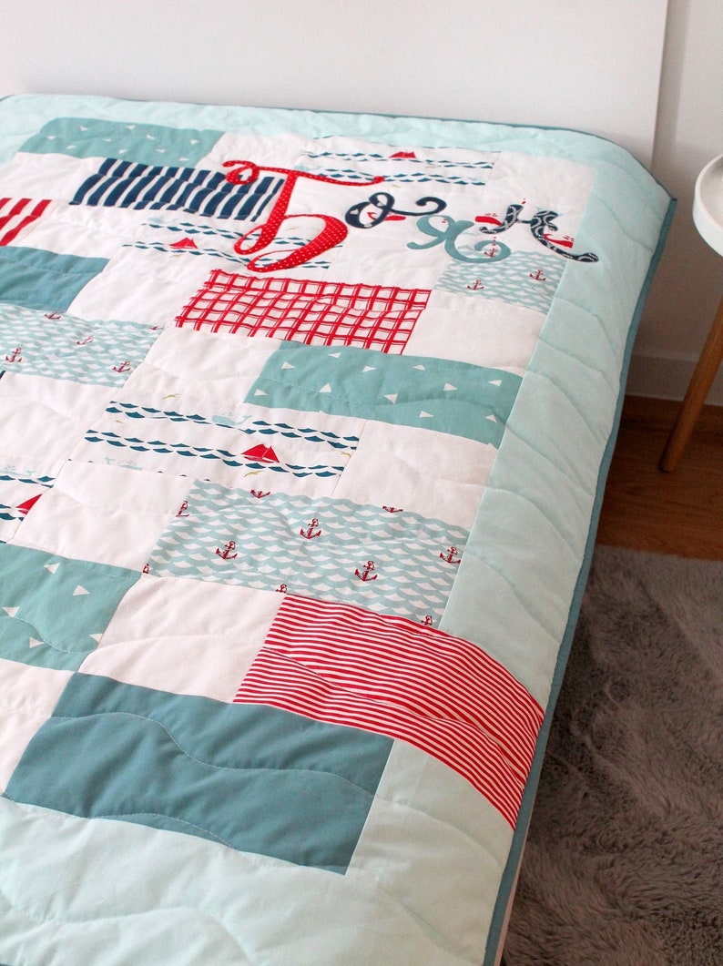 May include: A blue and white patchwork quilt with red and white stripes, anchors, and sailboats. The quilt has the name "J & J" embroidered on it.