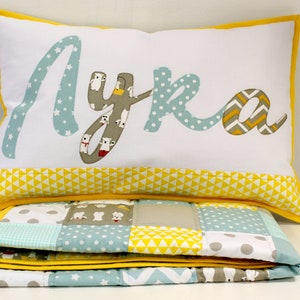 May include: A white pillow with a yellow border and the name "Lyra" spelled out in colorful fabric letters. The pillow is sitting on top of a folded quilt with a patchwork design in shades of blue, yellow, and gray.